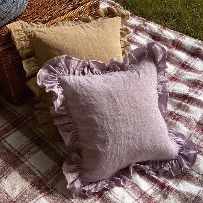 May include: Two square cushions with frilled edges. One cushion is a light lavender colour, and the other is a tan colour. The cushions are on a red and white plaid blanket, with a wicker basket in the background.