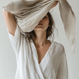 May include: A woman wearing a white wrap top and holding a beige scarf over her head. The scarf drapes down, partially obscuring her face. The clothing appears to be made of a soft, flowing fabric.