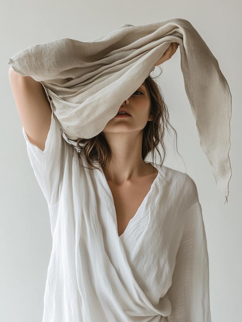 May include: A woman wearing a white wrap top and holding a beige scarf over her head. The scarf drapes down, partially obscuring her face. The clothing appears to be made of a soft, flowing fabric.