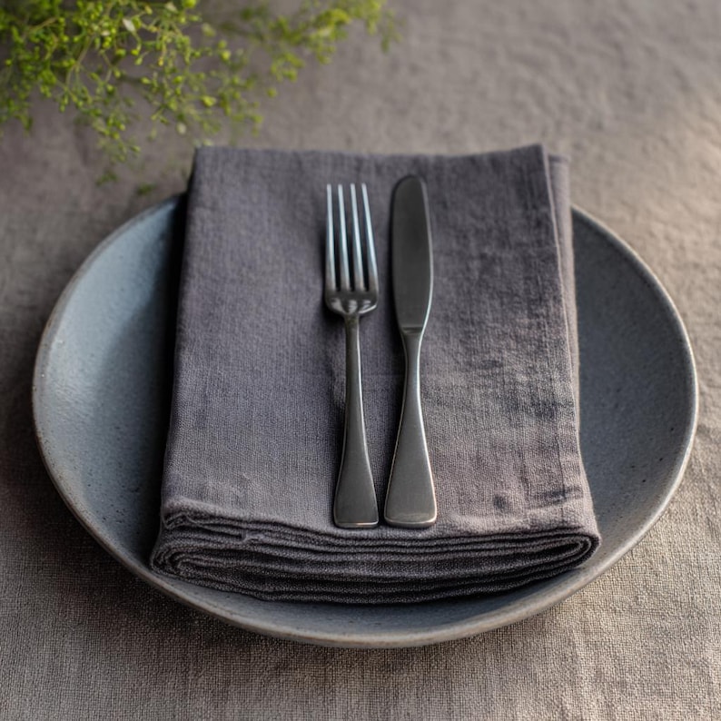 May include: A gray ceramic plate with a folded gray linen napkin, a fork, and a knife. The fork and knife are silver. The plate is on a gray tablecloth, with a sprig of green leaves in the background.