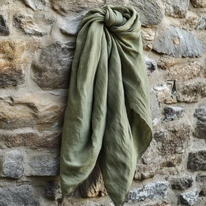 May include: A draped olive green scarf made of soft fabric. The scarf is knotted at the top and hangs down, with visible texture and a slightly frayed edge. The scarf is displayed against a stone wall.