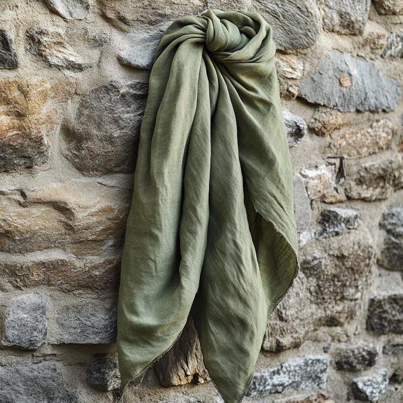 May include: A draped olive green scarf made of soft fabric. The scarf is knotted at the top and hangs down, with visible texture and a slightly frayed edge. The scarf is displayed against a stone wall.
