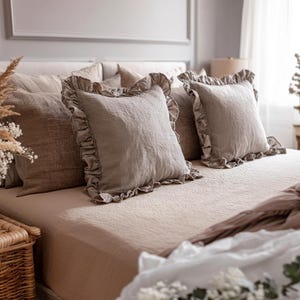 May include: A bed dressed in neutral-toned bedding, featuring several pillows. Two square pillows with ruffled edges are the focal point. The bed is covered with a textured, light brown sheet and a matching blanket. A wicker basket is visible in the foreground.