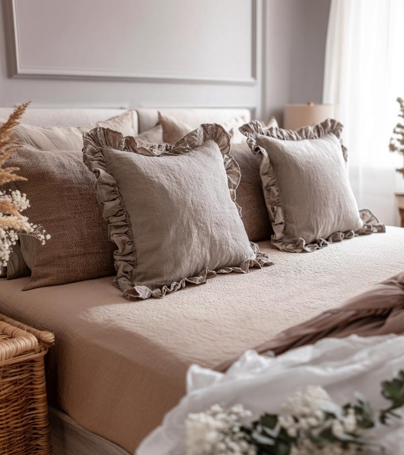 May include: A bed dressed in neutral-toned bedding, featuring several pillows. Two square pillows with ruffled edges are the focal point. The bed is covered with a textured, light brown sheet and a matching blanket. A wicker basket is visible in the foreground.
