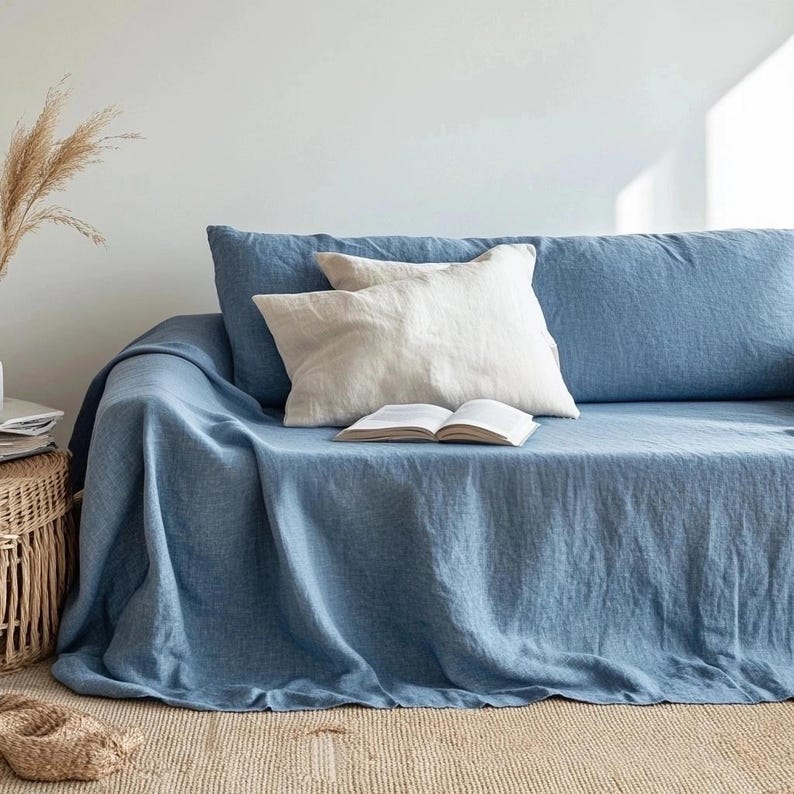 May include: A blue linen sofa cover draped over a sofa, with two pillows and an open book. The pillows are off-white. A woven basket and dried plants are visible in the background. The sofa is on a beige rug.