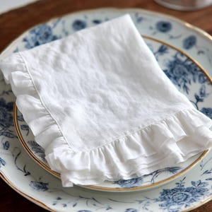 May include: A white linen napkin with ruffled edges, resting on a blue and white floral patterned plate with a gold rim. The napkin is folded and appears soft and slightly textured, suggesting a high-quality fabric.