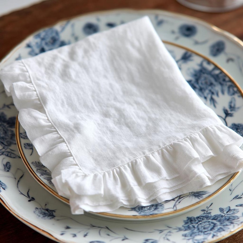 May include: A white linen napkin with ruffled edges, resting on a blue and white floral patterned plate with a gold rim. The napkin is folded and appears soft and slightly textured, suggesting a high-quality fabric.