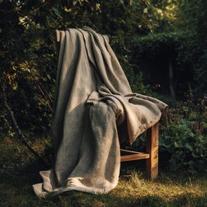 May include: A neutral-toned linen throw draped over a wooden chair, set outdoors. The fabric has a textured appearance, with folds and shadows adding depth. The chair is simple in design, with a natural wood finish. The scene is bathed in soft, natural light.