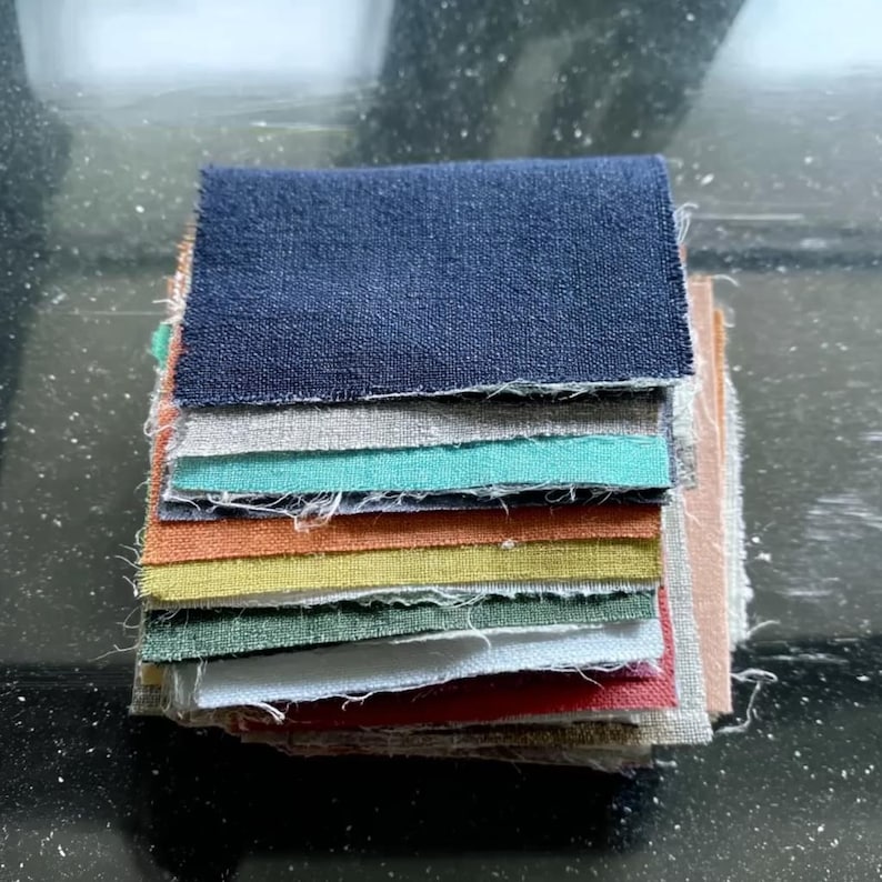 May include: A stack of fabric swatches in various colors, including navy blue, gray, teal, orange, yellow, and pink. The swatches are rectangular and have a textured appearance, likely linen or cotton. The edges are unfinished.