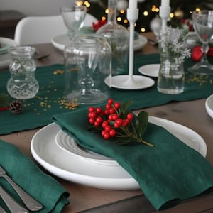 May include: A festive table setting with white plates, green napkins, and silverware. Red berries and greenery adorn the plates, with clear glassware and candle holders adding to the holiday ambiance. A dark green table runner completes the look.