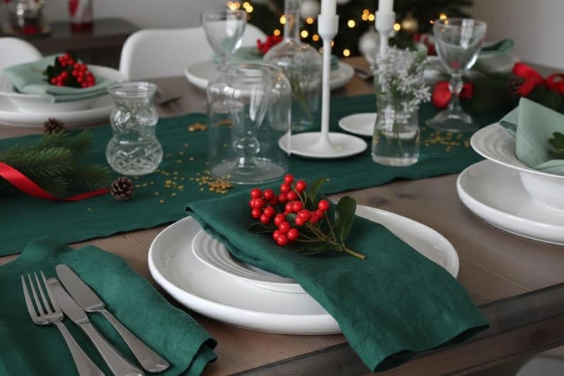 May include: A festive table setting with white plates, green napkins, and silverware. Red berries and greenery adorn the plates, with clear glassware and candle holders adding to the holiday ambiance. A dark green table runner completes the look.
