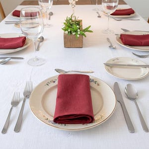 May include: A formal dining table setting with white tablecloth, floral patterned plates, and burgundy napkins. Silverware, water glasses, and a small wooden box with greenery complete the elegant arrangement. Ideal for a special occasion.