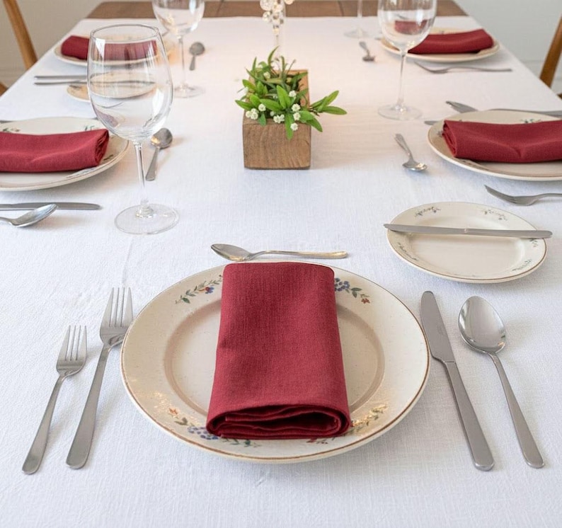 May include: A formal dining table setting with white tablecloth, floral patterned plates, and burgundy napkins. Silverware, water glasses, and a small wooden box with greenery complete the elegant arrangement. Ideal for a special occasion.