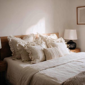 May include: A bed featuring a light-coloured wooden headboard and a selection of cream-coloured pillows. The pillows include ruffled and textured designs, arranged on a matching cream-coloured duvet. A black lamp with a white shade sits on a wooden bedside table.