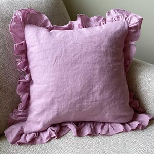 May include: A square, lilac-coloured cushion with a frilled edge. The cushion is made of a soft, textured fabric and sits on a patterned surface. The frilled trim adds a decorative touch to the cushion.