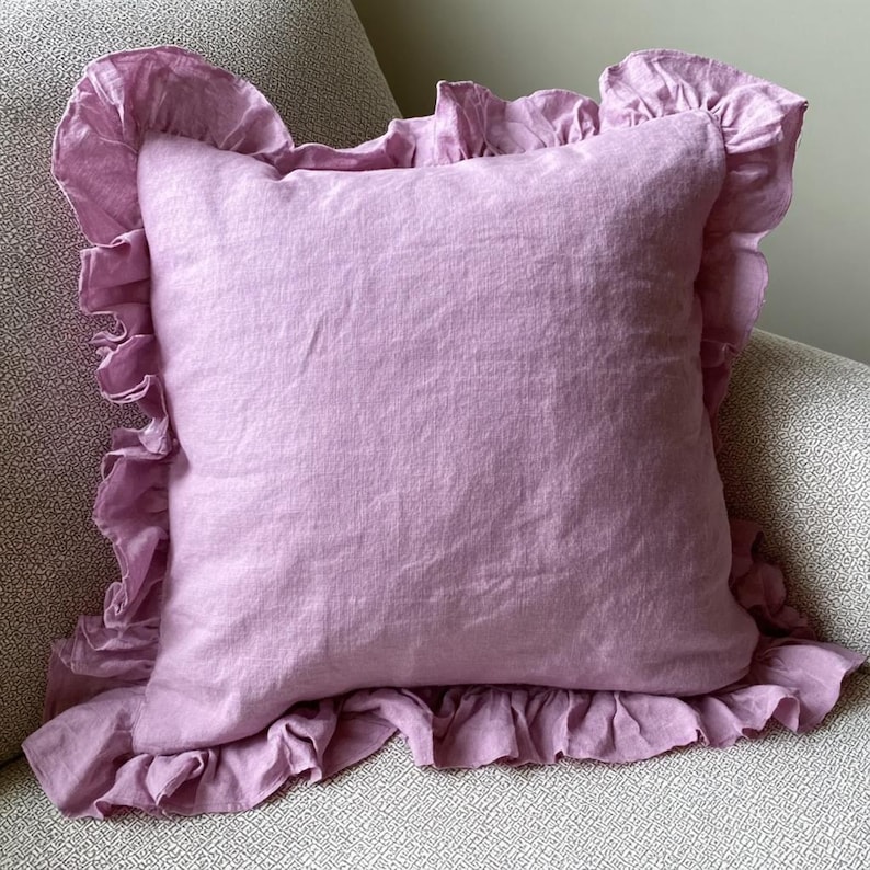 May include: A square, lilac-coloured cushion with a frilled edge. The cushion is made of a soft, textured fabric and sits on a patterned surface. The frilled trim adds a decorative touch to the cushion.
