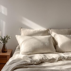 Op de afbeelding: Een bed met cr&egrave;mekleurig linnen beddengoed, inclusief kussens en een dekbed. De kussens zijn rechthoekig en op het bed geplaatst. Een kleine vaas met groen staat op een houten nachtkastje links van het bed. De kamer wordt verlicht door natuurlijk licht.