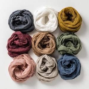 May include: Nine linen scarves in various colors, including navy, white, mustard yellow, burgundy, tan, olive green, blush pink, taupe, and denim blue. The scarves are arranged in a grid pattern on a white surface, showcasing their soft, textured appearance.