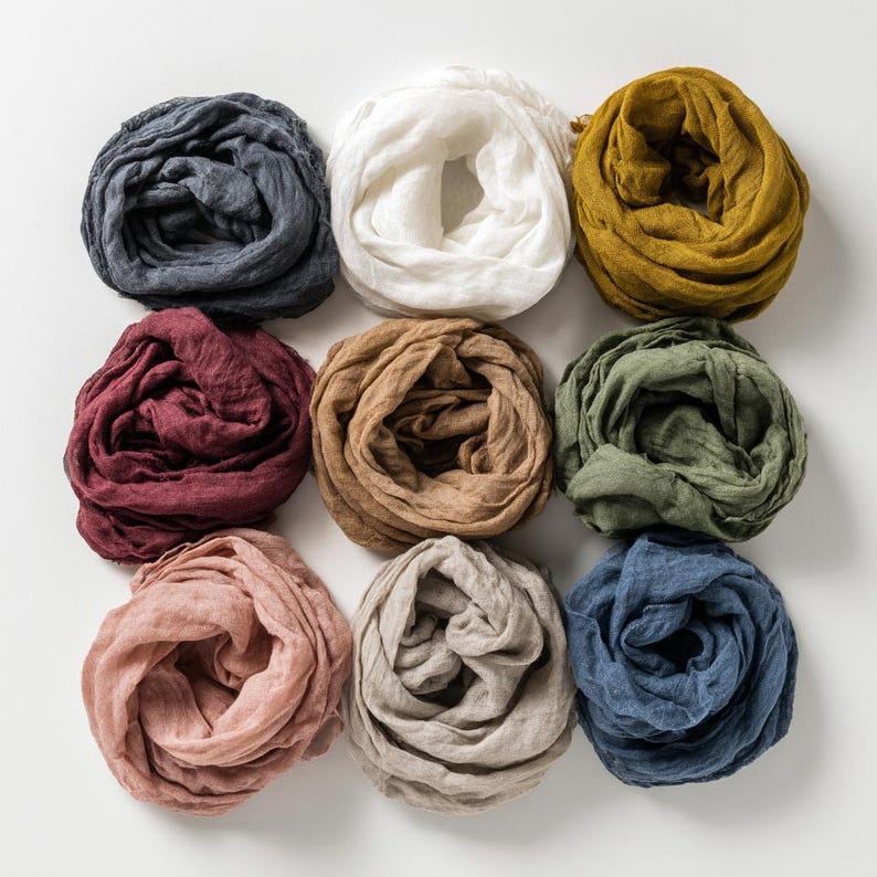 May include: Nine linen scarves in various colors, including navy, white, mustard yellow, burgundy, tan, olive green, blush pink, taupe, and denim blue. The scarves are arranged in a grid pattern on a white surface, showcasing their soft, textured appearance.