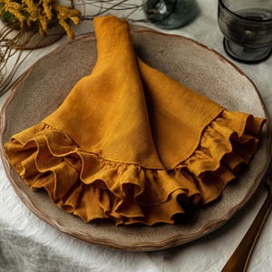 May include: A folded, mustard-yellow linen napkin with a ruffled edge, resting on a textured, round, ceramic plate. The napkin is the focal point, with soft folds and a decorative trim. The plate is a neutral color, and the background is blurred.