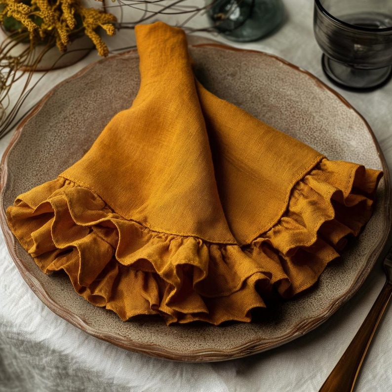May include: A folded, mustard-yellow linen napkin with a ruffled edge, resting on a textured, round, ceramic plate. The napkin is the focal point, with soft folds and a decorative trim. The plate is a neutral color, and the background is blurred.