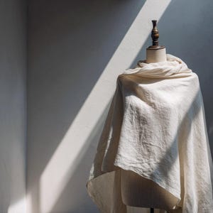 May include: A cream-colored linen shawl draped over a mannequin. The fabric has a soft, textured appearance. The mannequin is set against a white wall with a beam of sunlight.