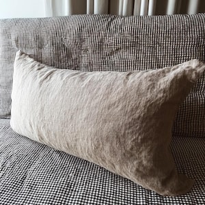 May include: A rectangular, light brown linen pillow rests on a black and white checkered patterned surface. The pillow has a textured appearance and is positioned against a checkered headboard.