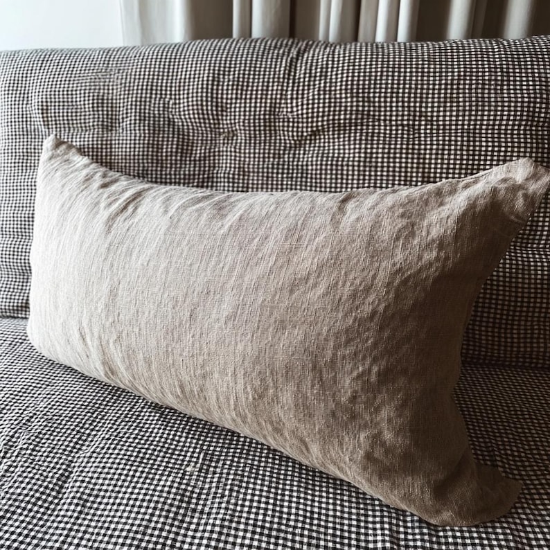 May include: A rectangular, light brown linen pillow rests on a black and white checkered patterned surface. The pillow has a textured appearance and is positioned against a checkered headboard.