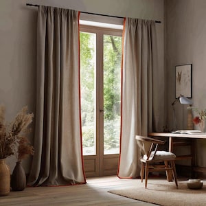 May include: Beige linen curtains with a vibrant orange trim frame a large window. The curtains are hung on a dark rod, creating a warm and inviting atmosphere. The room features a wooden desk, chair, and decorative vases.