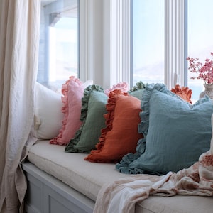 May include: A selection of colourful throw cushions with frilled edges, in shades of pink, green, orange, and blue, arranged on a window seat. A white bolster cushion and a patterned throw are also present. The window seat cushion is a neutral colour.