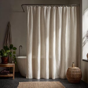 May include: A neutral-toned linen shower curtain hangs in a bathroom. The curtain is paired with a woven side table, a jute bath mat, and a white bathtub. A potted plant and a towel are also visible.