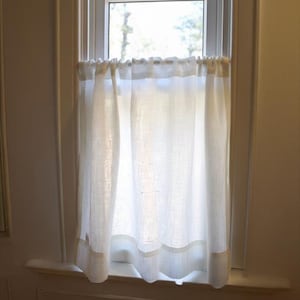 May include: A sheer, off-white linen curtain hangs in a window. The curtain has a simple design with a band of lighter fabric at the bottom. The window frame is white, and the background shows a glimpse of the outdoors.