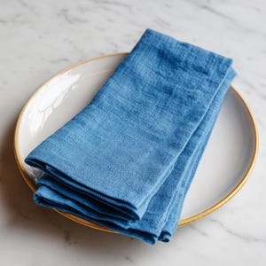 May include: A stack of folded blue linen napkins rests on a white plate with a gold rim. The napkins are a solid, light blue color, and the plate is set against a marble surface.
