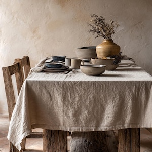 May include: A rustic dining table set for a meal. The table is covered with a beige linen tablecloth and set with stoneware bowls, plates, and mugs. A vase with dried flowers sits in the center. The table is made of weathered wood.