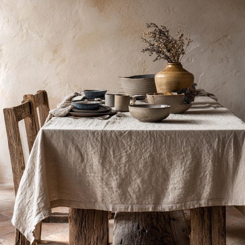 May include: A rustic dining table set for a meal. The table is covered with a beige linen tablecloth and set with stoneware bowls, plates, and mugs. A vase with dried flowers sits in the center. The table is made of weathered wood.