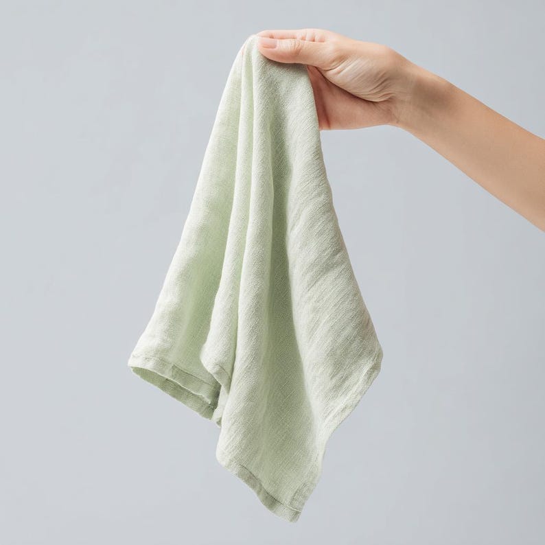 May include: A light green linen hand towel, held up against a light gray background. The towel is soft and textured, with visible woven threads. The edges are neatly hemmed, and the fabric drapes gently.