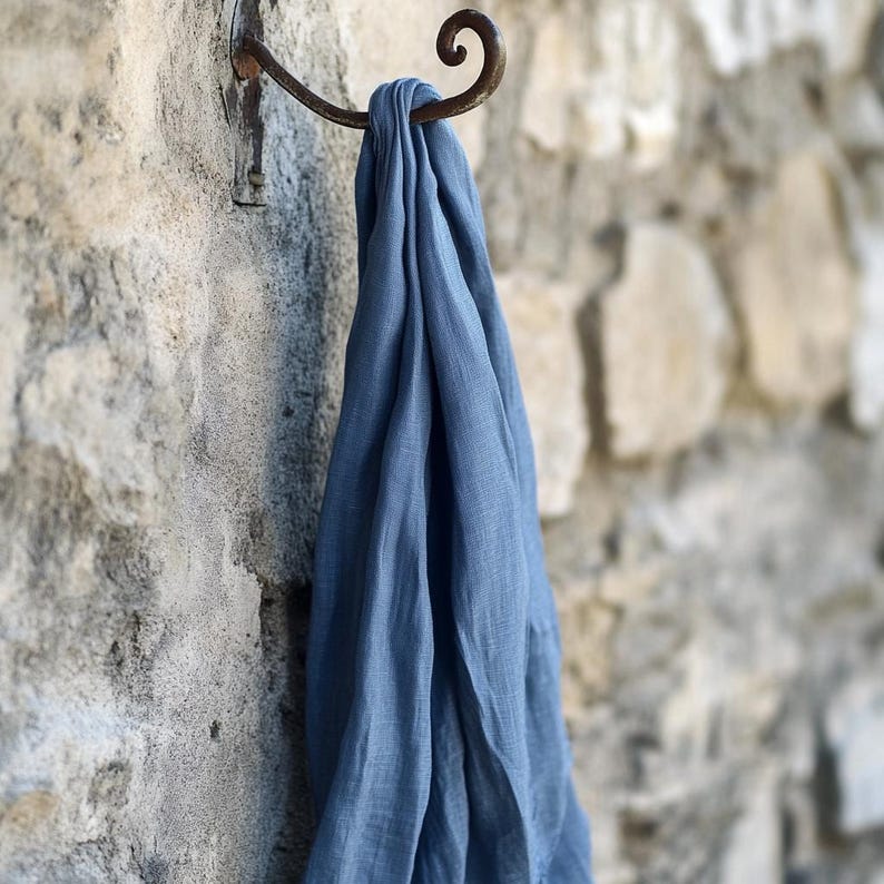 May include: A dusty blue scarf hangs from a rustic, wrought-iron hook on a textured stone wall. The scarf is made of a lightweight fabric and drapes in soft folds. The hook is mounted on a wall made of light-colored stones.