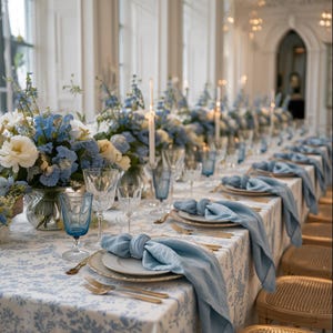 May include: A long dining table set for a formal event with a white and blue floral tablecloth. The table features blue linen napkins, clear crystal glassware, blue tinted water glasses, gold cutlery, and large floral centrepieces with blue flowers.