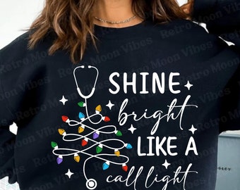 May include: Black sweatshirt with a white graphic that says "SHINE bright LIKE A call light." The design includes a stethoscope and a Christmas tree made of colorful lights and stars.