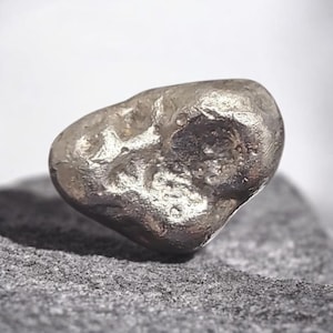 Op de afbeelding: Een kleine, onregelmatige metalen klomp met een reflecterend, zilverkleurig oppervlak. Het object heeft een getextureerde, oneffen buitenkant met diverse inkepingen en gladde, afgeronde randen, wat zorgt voor een organische en solide uitstraling.