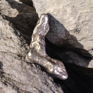 Puede incluir: Un objeto metálico en forma de L con una superficie texturizada e irregular y un acabado plateado reflectante. La pieza presenta contornos irregulares y un aspecto poroso y orgánico, descansando sobre un fondo de piedra natural gris.