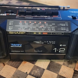 Könnte beinhalten: Ein schwarzer Sharp Kassettenspieler mit silberner Frontplatte und zwei Lautsprechern. Der Spieler hat ein digitales Display und ein Kassettendeck mit einem Doppelmechanismus. Der Text "Twin Mechanism Cassette Play" ist auf dem Spieler aufgedruckt.