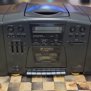 May include: A black Sansui stereo cassette deck with a silver faceplate and two speakers. The cassette deck has a digital display and a variety of buttons and knobs. The speakers are black and have a circular shape.