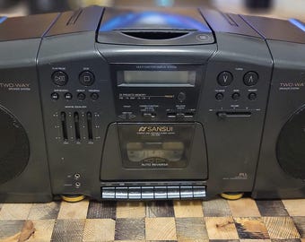 May include: A black Sansui stereo cassette deck with a silver faceplate and two speakers. The cassette deck has a digital display and a variety of buttons and knobs. The speakers are black and have a circular shape.