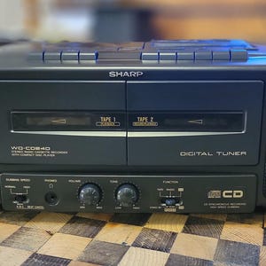 Può includere: Un sistema stereo Sharp nero con un registratore a cassette, un lettore CD e un sintonizzatore digitale. Lo stereo ha una placca frontale argentata con la parola "Sharp" in lettere bianche. Il registratore a cassette ha due slot per cassette etichettati "Tape 1" e "Tape 2". Il lettore CD ha una placca frontale argentata con la parola "CD" in lettere bianche. Il sintonizzatore digitale ha una placca frontale argentata con la parola "Digital Tuner" in lettere bianche.