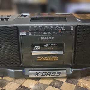 Könnte beinhalten: Ein schwarzer Kassettenspieler mit silberner Frontplatte. Der Spieler hat zwei Kassettendecks, einen Tuner und einen grafischen Equalizer. Der Spieler hat einen silbernen X-Bass-Knopf und den Text "Twincam Double Cassette" auf der Vorderseite.
