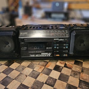 Puede incluir: Boombox negro Sharp con altavoces a cada lado. El boombox tiene un reproductor de casetes, un dial de radio y varios botones de control. Las palabras "Twin Mechanism W" son visibles. Sobre una superficie de madera a cuadros.