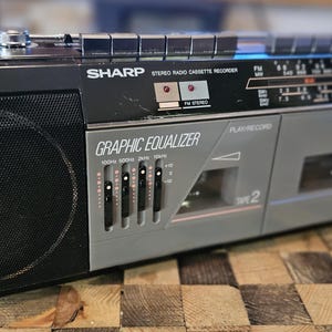 以下が含まれることがあります： ヴィンテージの黒とグレーのSharpステレオラジオカセットレコーダー。ブームボックスは、グラフィックイコライザー、カセットデッキ、ラジオダイヤルを備えています。スピーカーは黒いメッシュカバーが付いています。