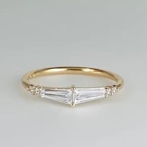 May include: A delicate gold ring with a central cluster of baguette-cut diamonds. The ring has a slender band and is embellished with smaller round diamonds on either side of the central stones. The ring is set against a plain white background.