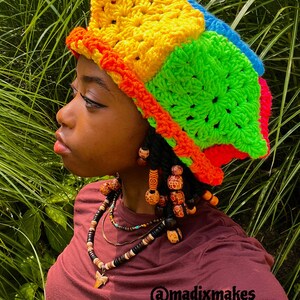 May include: A vibrant, handmade crochet hat in a patchwork design. The hat features bright colors including yellow, green, blue, and red. The image also shows a maroon shirt and beaded jewelry. The text "@madixmakes" is visible.